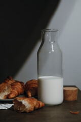 Glass decanter with milk and plate with fresh croissants, grey background. Healthy breakfast concept. Selective focus, close up.
