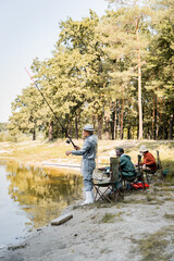 Asian man fishing near interracial friends on chairs on lake coast