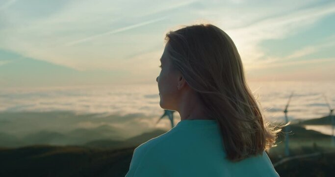 Soft focus on profile of beautiful young woman stand on top of hill looking at wind turbines at sunset. Concept mindfulness and renewable energy. Peaceful new generation focused on sustainability 