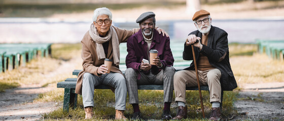 Multiethnic senior men with paper cups, smartphone and walking cane sitting on bench in park, banner