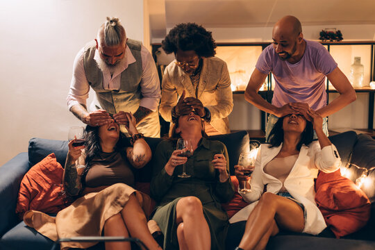 dinner with friends - men playing to cover the eyes of women sitting on the sofa on chatting - festive reunions and good mood