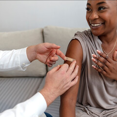 Happy patient laughs after vaccination while her doctor puts a band-aid on her arm. African woman...