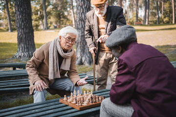 Fototapeta na wymiar Senior interracial friends playing chess near friend pointing with finger in park