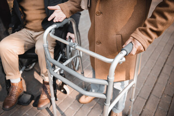 Obraz premium Cropped view of senior man with walking frame standing near friend in wheelchair outdoors