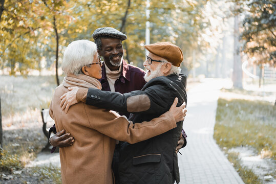 Cheerful elderly man hugging asian and african american friends in autumn park