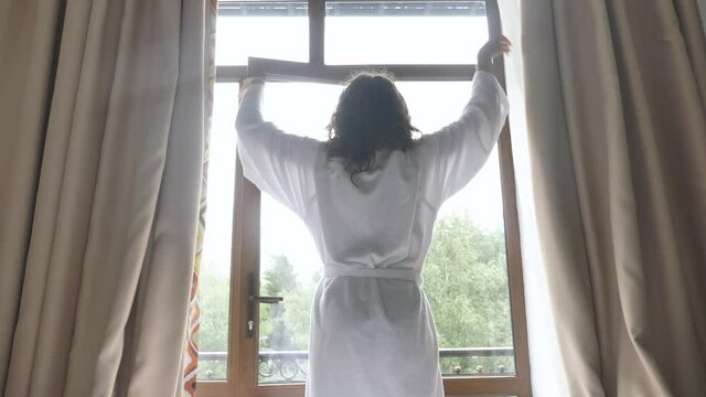 Curly haired young woman in white bathrobe opens light brown curtains and stretches arms near balcony door in morning backside view