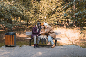 Positive interracial elderly men hugging on bench in park