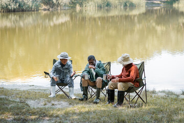 Positive interracial senior men in fishing outfit holding thermo cups while spending time outdoors