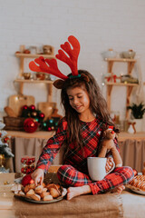 cute little brunette girl in checkered red pajamas with reindeer horns on her head is eating a Christmas cake and drinking tea from a white mug in a beautifully decorated kitchen. lifestyle, hygge © КРИСТИНА Игумнова