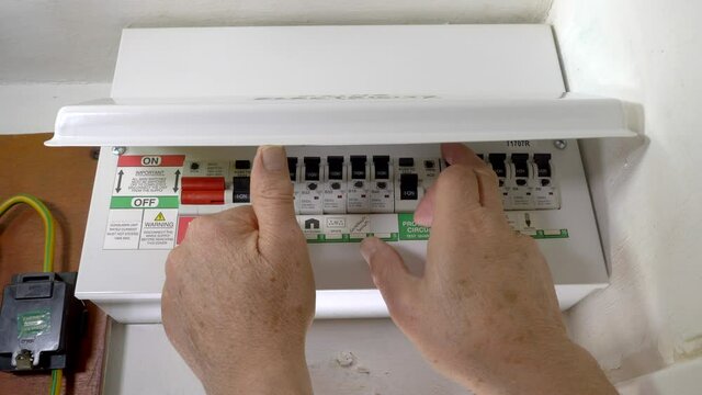 An electrician&rsquo;s hands raising the cover of a domestic mains electricity fuse box / consumer unit, attached to an interior wall, then switching off the RCD protected circuit for a shower.