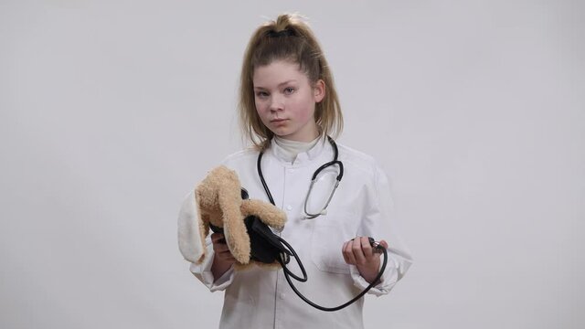 Focused Caucasian Girl In Nurse Gown Measuring Toy Blood Pressure With Sphygmomanometer And Looking At Camera Smiling. Confident Child Practicing Dream Job Posing At White Background