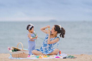 Mother and baby are happy on a sunny day at the beach where they sit and have a picnic together in a fun mood and learn together by playing with a toy camera.