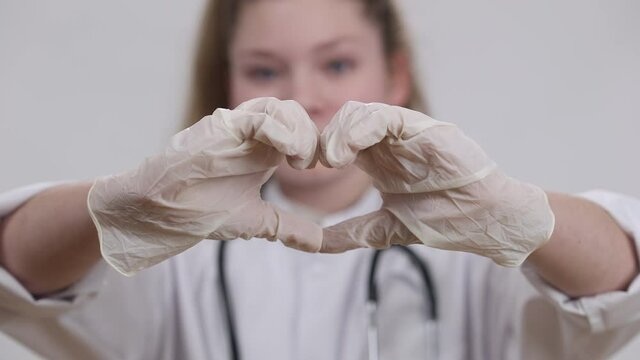 Close-up little female hands in medical gloves showing heart shape with blurred smiling pretty kid looking at camera at background. Confident Caucasian child choosing medical profession as dream job