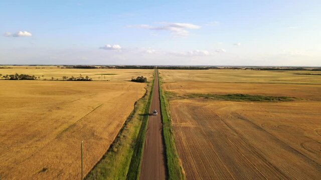 Silver car driving along straight endless dirt road swirling up dust during sunset in Canada's countryside. 4k tracking aerial view from high above.