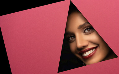 Face of a young beautiful girl with a bright make-up and with plump red lips peeks into a hole in pink paper.