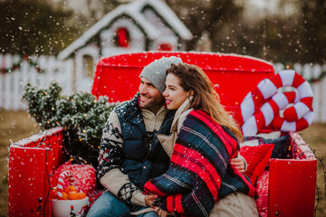 Handsome young man and pretty curly haired woman in winter clothes posing in red Christmas car with decor against white house and fence. Focus is at the woman. Snowing. Holiday concept.