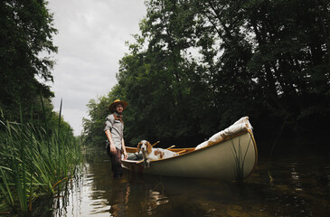 Young man stands in shallow river next to his canoe with a dog