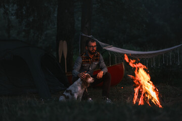Person sits next to his dog by the bonfire, camping with canoe in beautiful nature