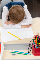 A cute first grader boy in a school uniform at home during a pandemic fell asleep doing homework at a desk with books and pencils. Selective focus. Close-up. Portrait