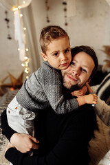 Portrait of happy father hugging with little son, looking at the camera, smiling. Loving parent spending winter holidays with his child at home, enjoying time together, fatherhood concept