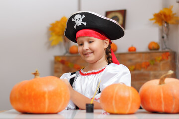 Celebrating Halloween at home. A little girl dressed as a pirate sits at the table. The child prepares for the party and draws on the pumpkin. - obrazy, fototapety, plakaty