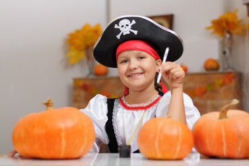 Celebrating Halloween at home. A little girl dressed as a pirate sits at the table and draws on a pumpkin. The child looks into the camera. - obrazy, fototapety, plakaty