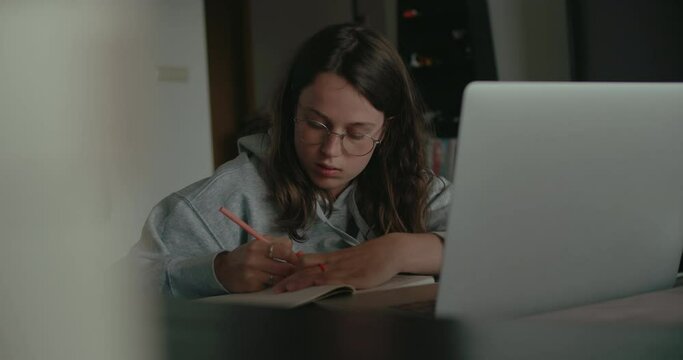 Cinematic shot of young student doing homework at home, work on school project or working from home in cosy living room. Comfortable home office setup for study or work. Make notes in notebook from