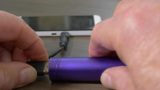 Closeup POV Shot Of A Man’s Hands Putting Down A Mobile Phone With Attached Lead / Cable / Cord, And Plugging It In To The USB Port Of A Portable Charger / Battery Power Bank.