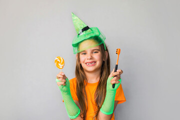 Happy Halloween. St. Patrick's Day.  Blonde girl kid in a witch costume with a candy and a toothbrush in her hands. Place for text, place for copying. Dental hygiene, children's health concept.