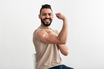 Young Arab man with adhesive bandage getting vaccinated against covid-19, demonstrating his muscles on white background