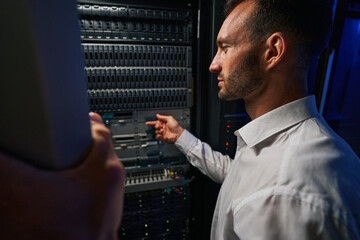 Bearded busy server administrator in white shirt examining work of supercomputer in tech room