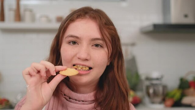 Portrait of young girl eat cookie and look delicious that she stay in kitchen and look at camera with happiness emotion in her house.