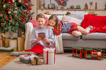 Two joyful sisters are sitting at home near the christmas tree and looking at tablet monitor