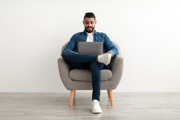 Young Arab man in casual wear working online, sitting in armchair and using laptop against white studio wall