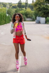 full length of trendy african american woman skating on roller skates, drinking orange juice and holding cellphone