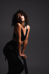 Fototapeta premium brunette african american woman in trendy outfit posing on wooden chair on dark grey