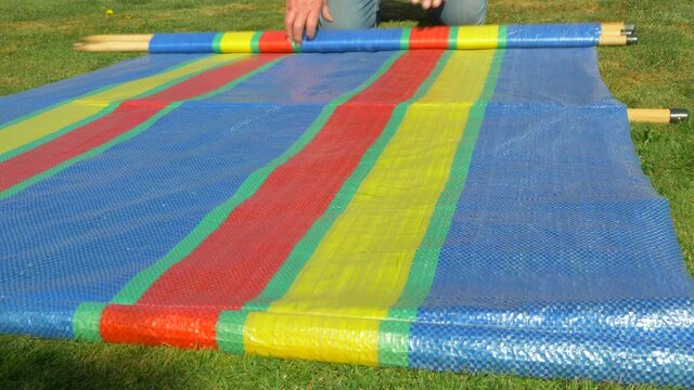 Closeup POV shot at ground level of a man&rsquo;s hands unrolling a colorful camping windbreak / windscreen and wooden poles on a piece of grass.