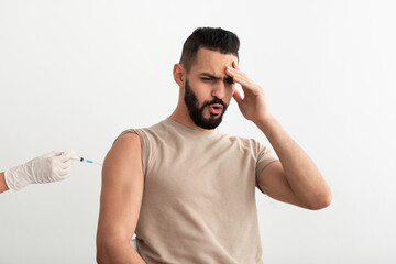 Young Arab guy feeling scared of covid vaccination, taking coronavirus vaccine shot on white studio...