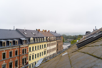 Fototapeta premium Colorful rooftops of residential buildings with landscape aerial panoramic view to green foggy forest or park on cloudy summer or autumn day. View from window or balcony in Dresden, Germany