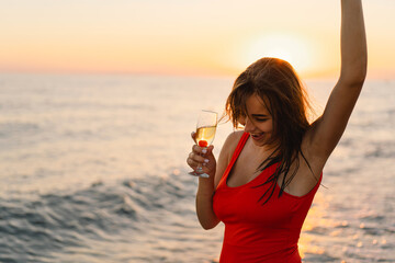 Young beautiful woman in a red swimsuit and in a Santa hat with a glass of champagne in her hands walk on the beach. Woman celebrate Christmas and new year at tropics. New Year concept.