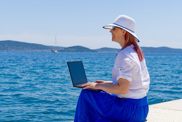 Work from anywhere. Side view of young woman, female freelancer in straw hat working on laptop...
