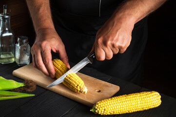 Professional chef cuts fresh corn with a knife. Boiled corn is a great diet for breakfast or lunch. Working environment on the restaurant kitchen table.