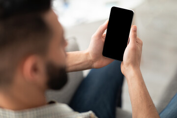 Unrecognizable young Arab man using smartphone on sofa at home, mockup for app or website design