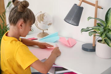 Cute little girl making paper plane at table in room