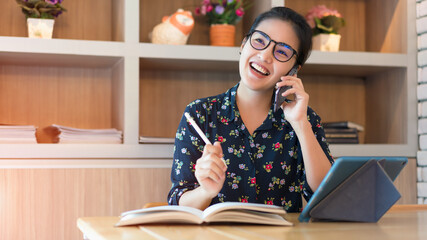 An Asian businesswoman in glasses sits at home with a laptop and a notebook on the side in her hand holding a pen and is using a mobile phone in soft light in the morning.