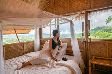 Young asian woman resting and looking at outside on the bed in wooden cottage among the mountain