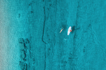 Man rowing on SUP board on aquamarine, blue sea water with coral in lagoon. Woman swimming near her boyfriend. Couple resting, doing sport, healthy recreation. Aerial top view paddleboard. Surfing - Powered by Adobe