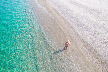 Young athletic woman in hat, swimsuit walking barefoot on coast of sea on the sand beach. Summer. View from above. Top view, copter