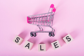 SALES - word on wooden cubes, on a pink background with a shopping trolley.