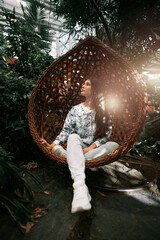 A young girl sits in a cocoon hanging chair. Brunette woman in the indoor urban jungle